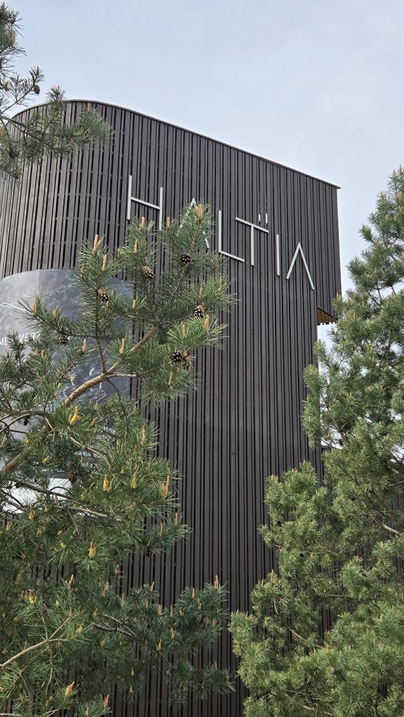 Rakennuksen julkisivu, jossa on pystysuuntainen puuverhous. Etualalla havupuita ja taustalla pilvinen taivas.